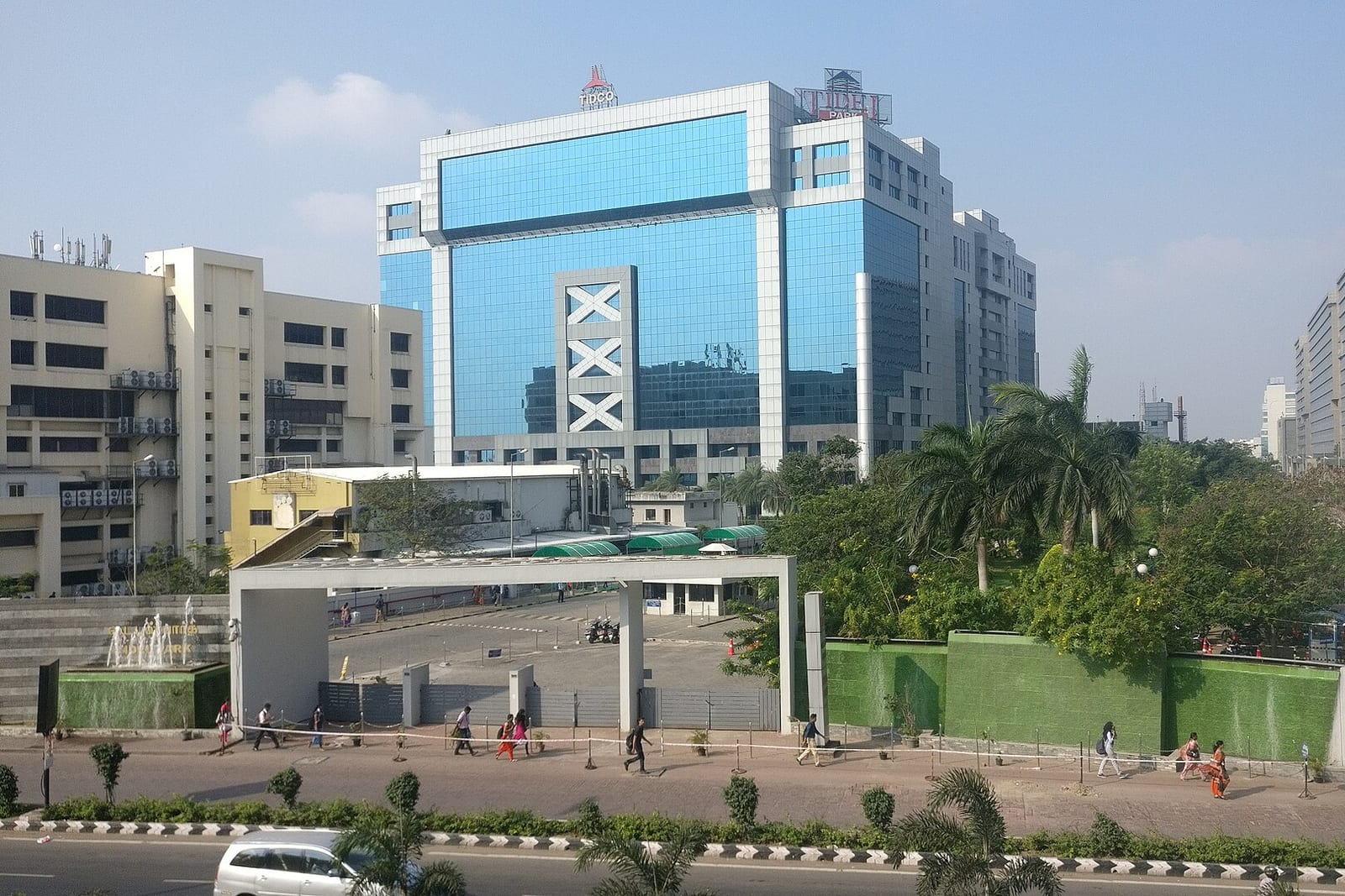 Blue-glass office buildings behind a gate, with palm trees and a road in front