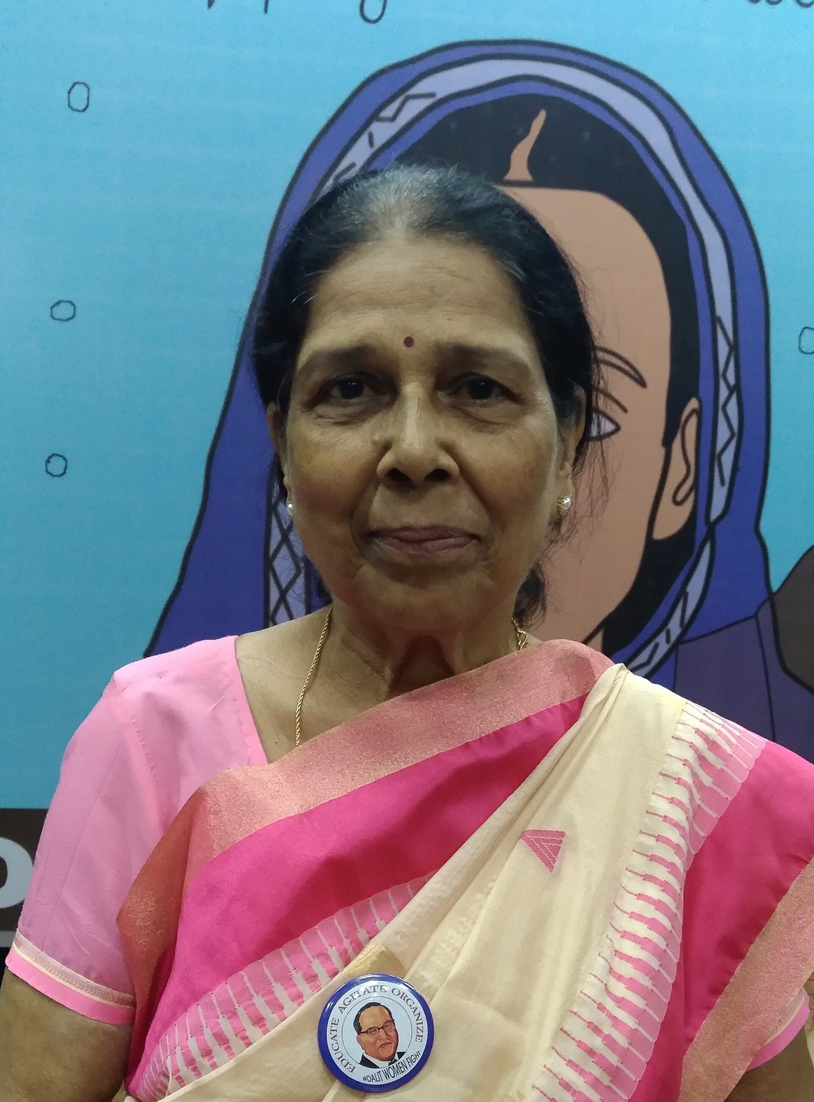 Urmila Pawar in a pink and cream sari, wearing a badge with a portrait of B.R. Ambedkar and the words Educate Agitate Organize #DalitWomenFight, in front of an illustrated poster