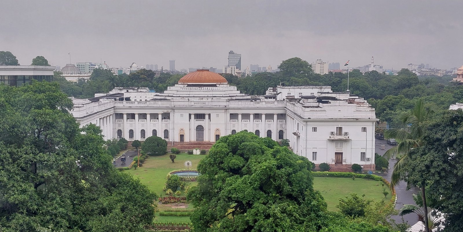 The white, colonnaded West Bengal Legislative Assembly building in Kolkata, with a copper dome, seen above trees under a grey sky